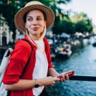Cheerful female with rucksack smiling and looking away while standing on quay near river with smartphone in hand during vacation
